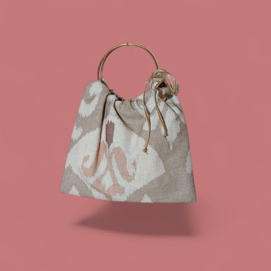 Bolso de tela beige con diseño abstracto y asa dorada, sobre fondo rosa.
